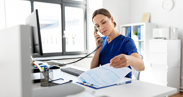 Eine Frau sitzt am Schreibtisch und blätter in Unterlagen während sie den Telefonhörer ans Ohr hält.