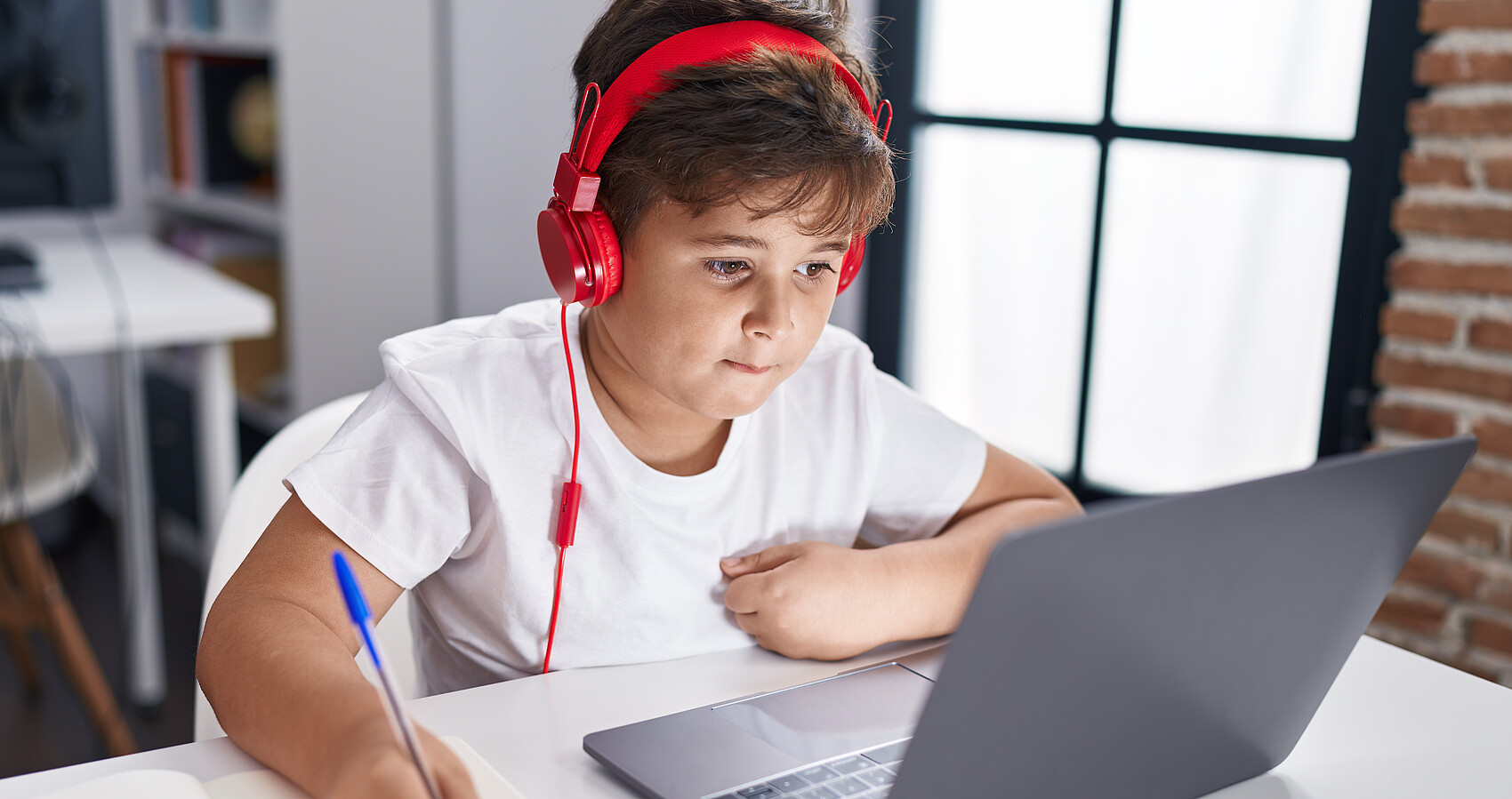 Schüler im Klassenzimmer vor seinem PC mit rotem Kopfhörer, der in ein Notizbuch schreibt
