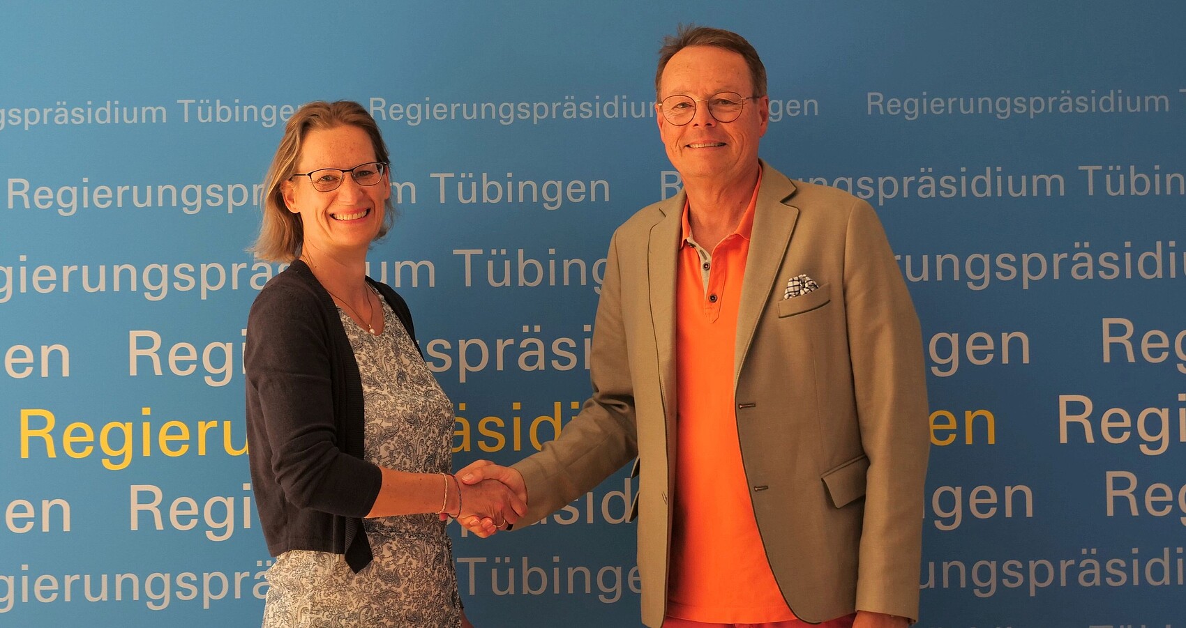 Das Foto zeigt Claudia Schneiderhan und Regierungspräsident Klaus Tappeser die sich die Hand reichen vor einer Plakatwand des RP Tübingen
