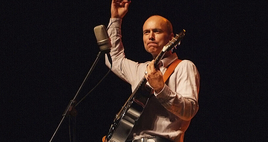 Das Bild zeigt einen glatzköpfigen Mann vor einem Mikrofon. Er hat eine Gitarre umhängen, die er mit der linken Hand hält, die rechte Hand streckt er nach oben. Der Hintergrund ist schwarz. 