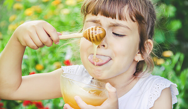 Ein Mädchen hält eine Schale mit Honig in der Hand und lässt mit einem Löffel Honig auf die Zunge fließen.