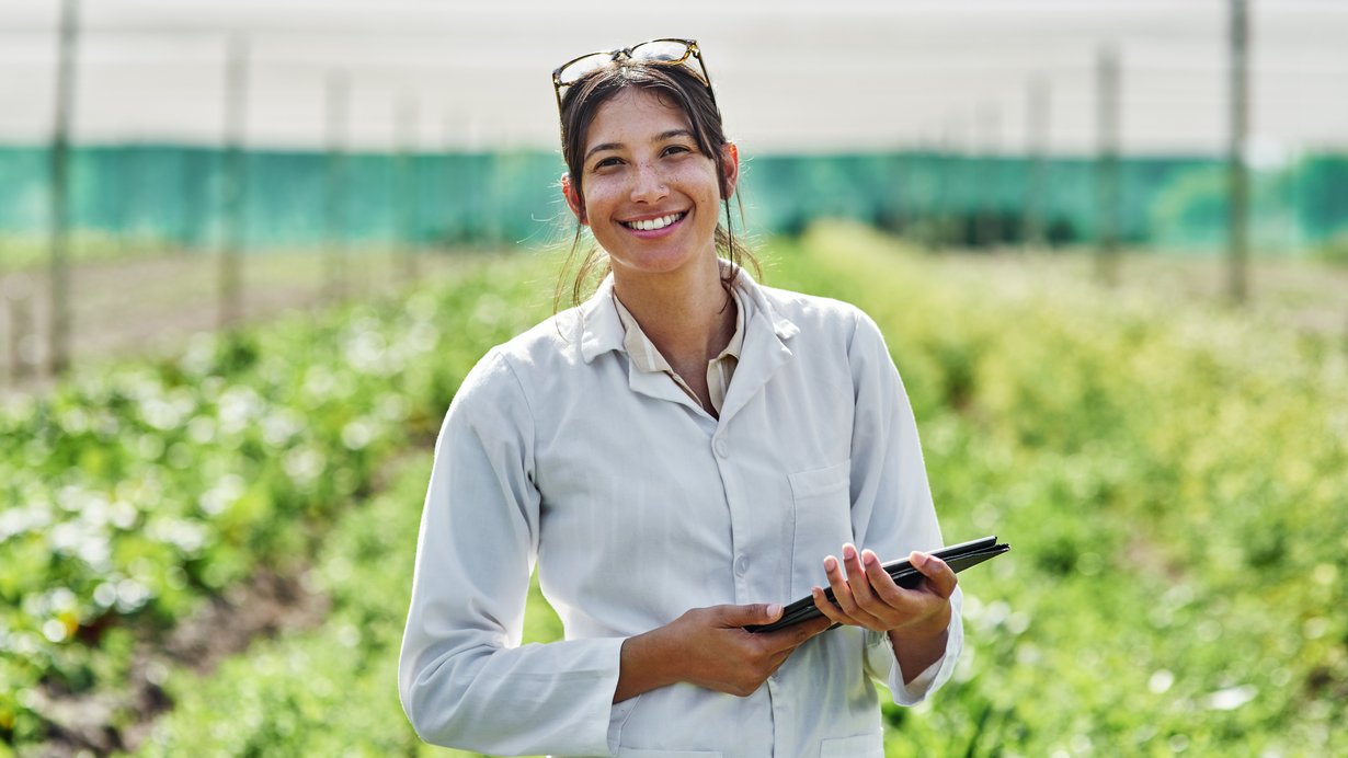 Eine Frau steht mit einem Laptop in der Hand auf dem Fled und lacht in die Kamera