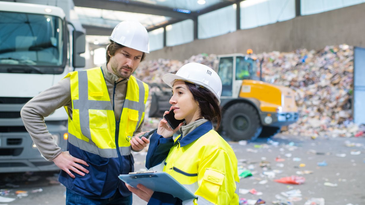 Weibliche und männliche Fachkraft für Kreislauf- und Abfallwirtschaft. Links dahinter ein Lkw, im Hintergrund rechts liegt recyclebares Material