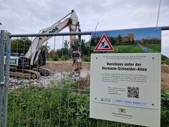 Informationsschild, welches an einem Bauzaun befestigt ist. Im Hintergrund arbeitet ein Bagger. Auf dem Informationsschild ist Ziel und Inhalt der Maßnahme zu lesen. Der Text des Schildes lautet: Hier führt der Landesbetrieb Gewässer eine vorbereitende Maßnahme für den Bau des Polders Bellenkopf-Rappenwört durch: Durchlass unter der Hermann-Schneider-Allee.