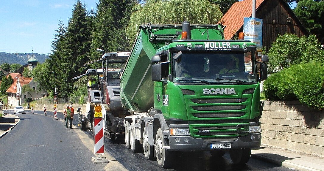 Ein grüner Lastwagen schüttet Asphalt in einen Aufbereiter