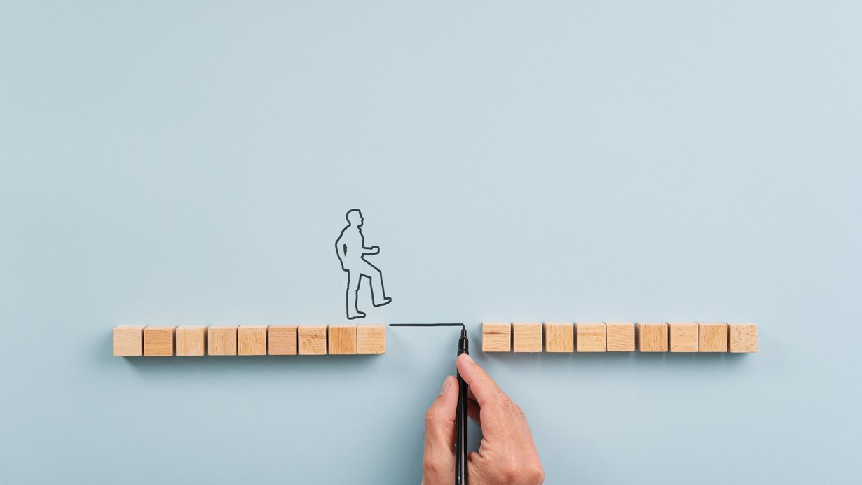 Holzwürfel bilden einen Weg mit Lücke, eine skizzierte Person überquert ein gezeichnetes Verbindungsstück zwischen den beiden Wegseiten.