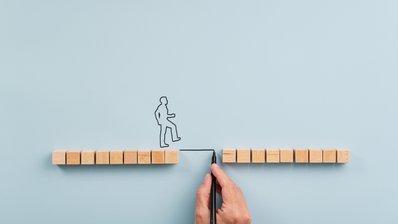 Holzwürfel bilden einen Weg mit Lücke, eine skizzierte Person überquert ein gezeichnetes Verbindungsstück zwischen den beiden Wegseiten.