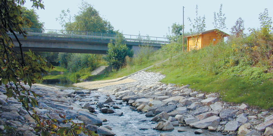 Zu erkennen ist ein Fluß, der wenig Wasser führt. Am rechten Ufer sieht man ein Häuschen aus Holz. Darin befinden sich die Geräte und Instrumente zur Pegelmessung.