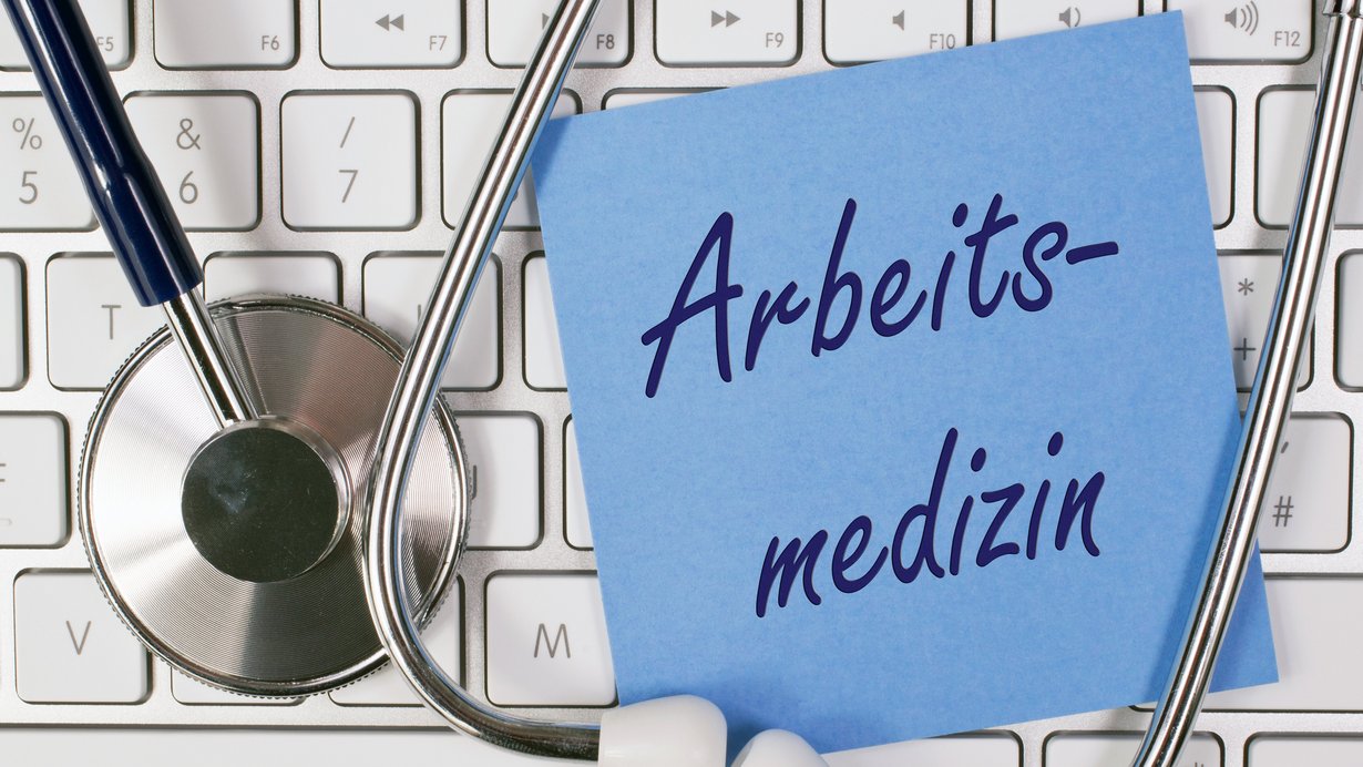 Bild zeigt Stethoskop mit Tastatur und einem Notizzettel mit der Aufschrift "Arbeitsmedizin"