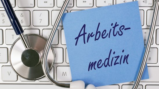 Bild zeigt Stethoskop mit Tastatur und einem Notizzettel mit der Aufschrift "Arbeitsmedizin"