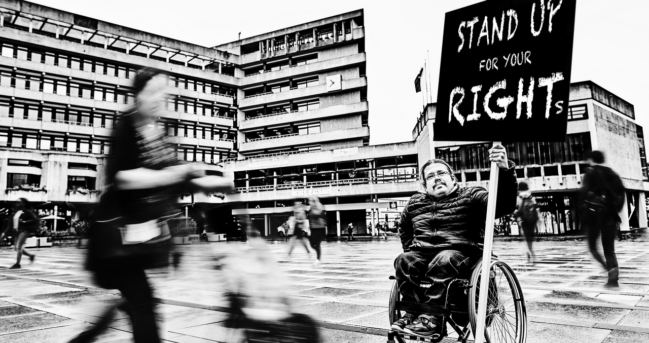 Auf einem Platz vor einem großen Gebäude hält ein Mann im Rollstuhl ein Schild hoch mit der Aufschrift "Stand up for your rights".