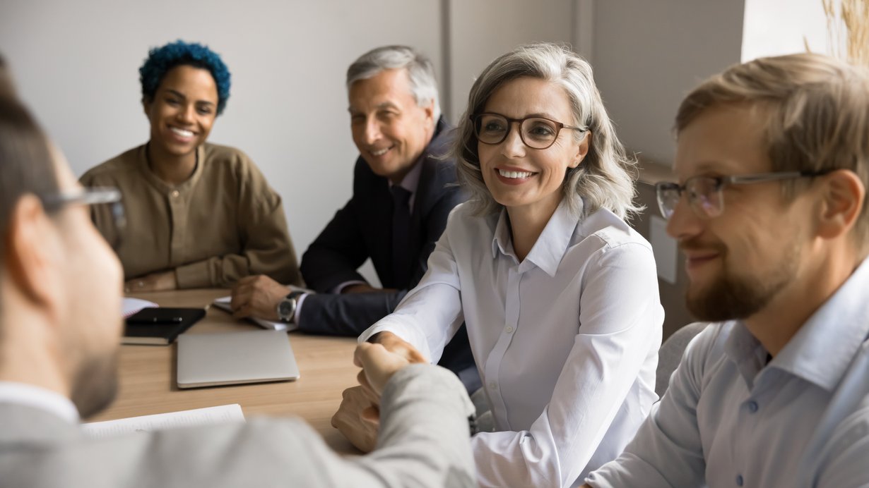 Lehrerinnen und Lehrer verschiedenen Alters sitzen am Tisch. Die ältere Kollegin schüttelt die Hand eines jungen Kollegen.