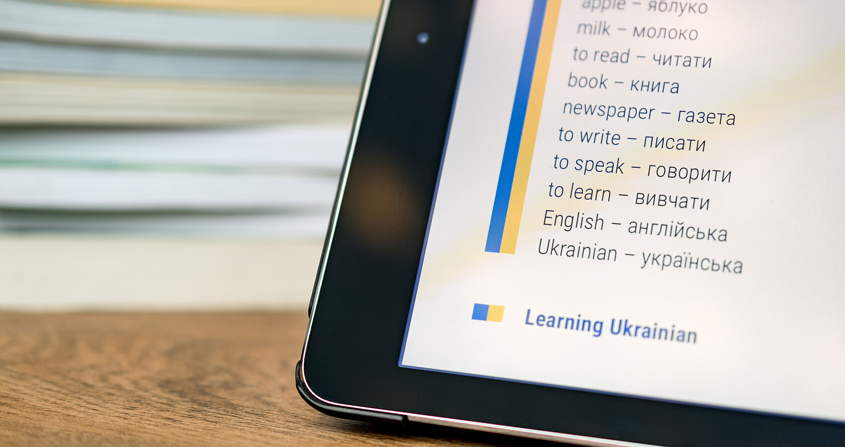 Eine Ecke eines Sprachcomputers wird im rechten Bildrand gezeigt. Darin sind englisch-ukrainische Vokabeln zu lesen. Im Hintergrund liegen Bücher aufgestapelt.
