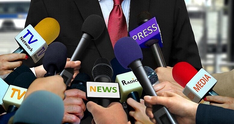 Vor einem verschwommenen Hintergrund sieht man den Oberkörper eines Mannes. Er trägt ein schwarzes Jacket über einem hellblauen Oberhemd und eine leicht glänzende, granatrote Krawatte. Ihm strecken sich elf Hände mit Mikrofonen unterschiedlicher Bauart und Farbe entgegen, an denen Schilder mit Aufschriften wie „TV Network“, „TV“, „News“, „Radio“, „PRESS“ und „Media“ befestigt sind.