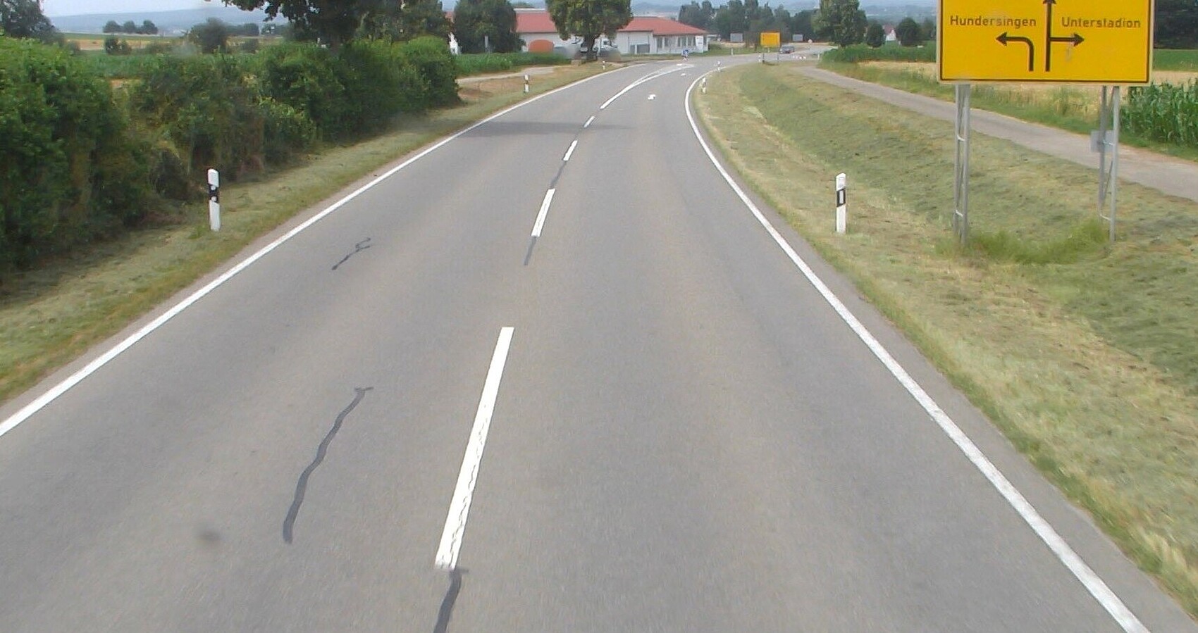 Das Foto zeigt eine einspurige Straße mit Fahrbahnmarkierung. Leitplanken. Links und rechts Grünstreifen und ein gelbes Straßenschild