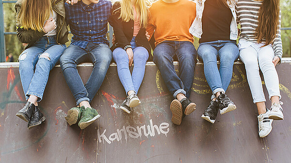 6 Kinder in bunter Kleidung auf einer Mauer sitzend, dabei sieht man nur den Unterkörper der Kinder, die ihre Beine baumeln lassen. Die Mauer trägt den Schriftzug „Karlsruhe“