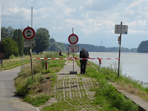 Das Foto ist entlang des Rheinhochwasserdamms aufgenommen. Der Blick geht von der Rheinbrücke in Richtung Hofgut Maxau. Man sieht, dass aufgrund des anstehenden Hochwassers der Damm mit Warnschildern und rot-weißem Band abgesperrt ist. Das Betreten ist verboten. Es besteht Lebensgefahr. Trotzdem sind mehrere Menschen auf dem Damm unterwegs und nutzen die dortigen Bänke für die Aussicht auf den Rhein bei Hochwasser. Ein Mann lehnt vorn an der Absperrung und hält sich daran, sie nicht zu überqueren. 