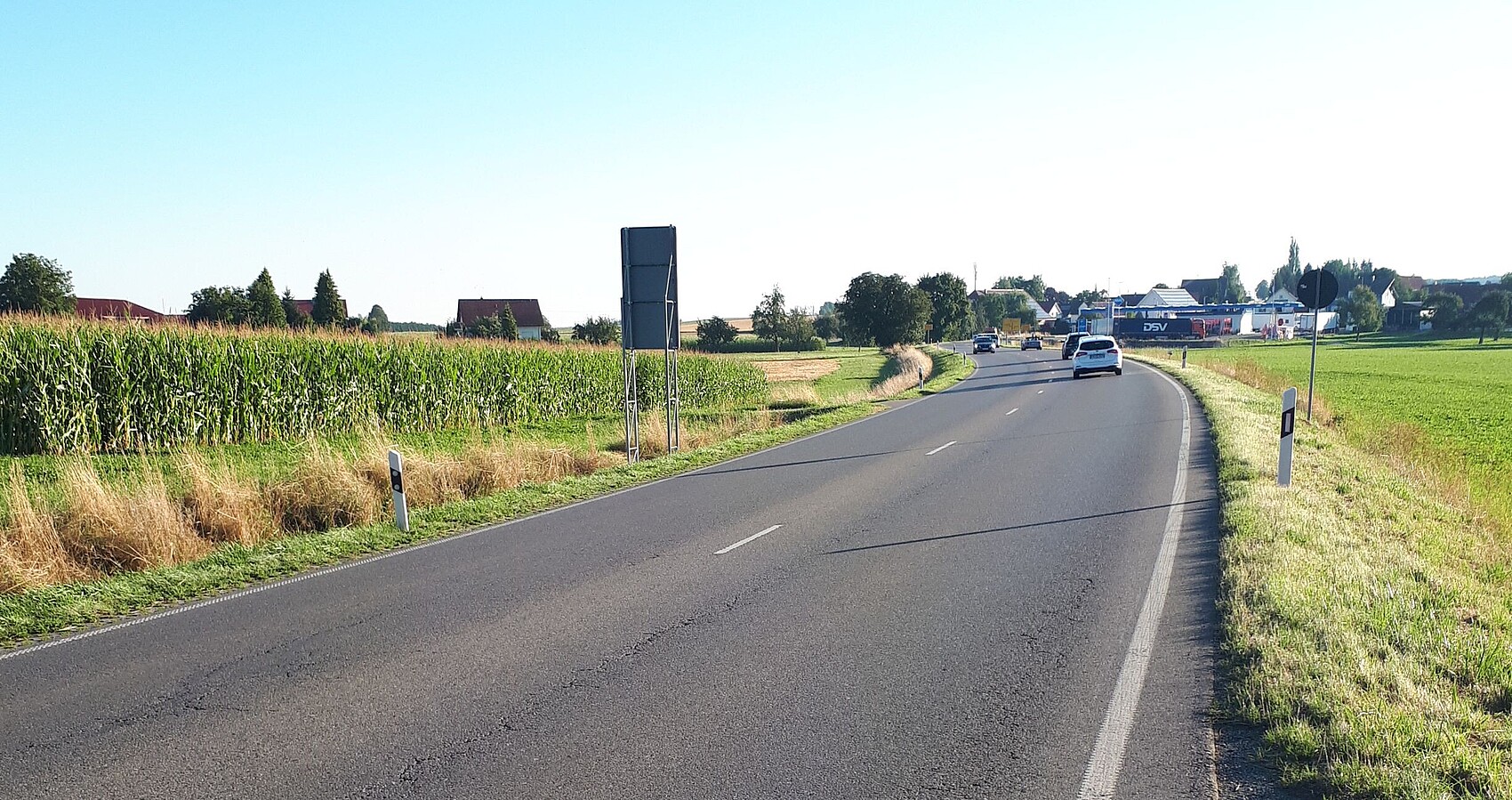 Das Bild zeigt die B 30. Links davon ein Maisfeld und rechts eine Wiese. Im Hintergrund sieht man mehrere Gebäude von Enzisreute. 