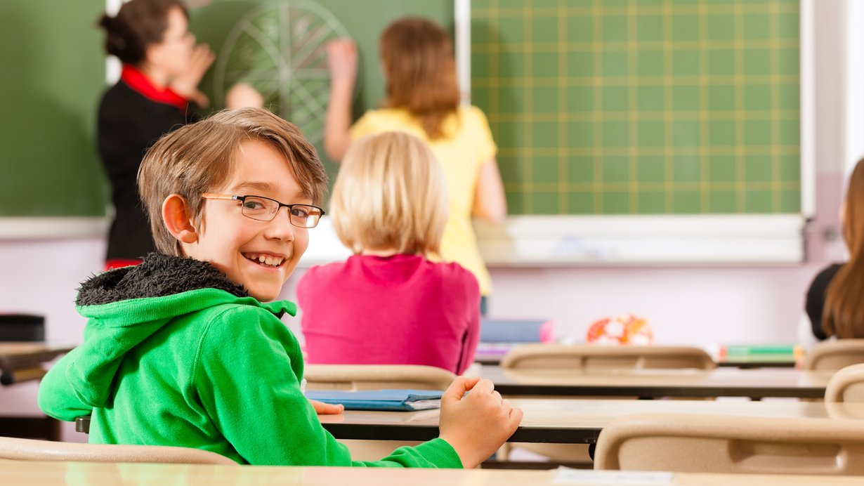 Ein Lehrerin unterrichtet Schüler in einer Schule. Ein Schüler blickt lachend in die Kamera.