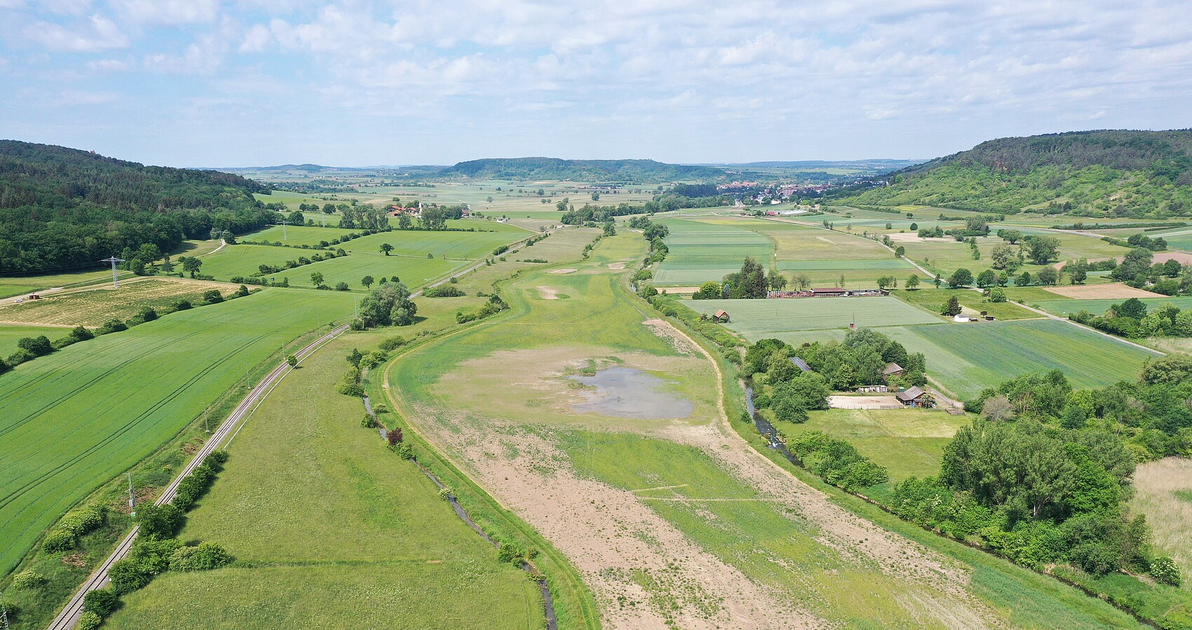 Wiedervernässte Ackerflächen im Ammertal; Bahnlinie links; links und rechts Wald; im Hintergrund blauer bewölkter Himmel