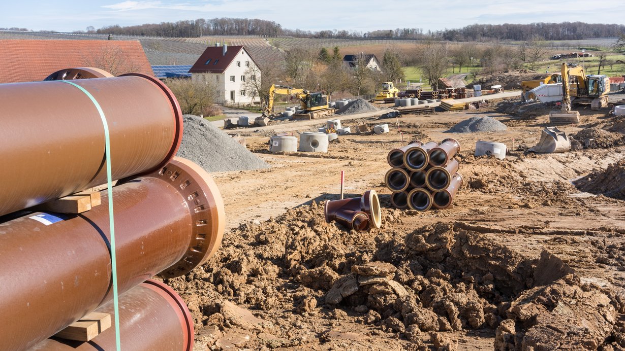 Im Vordergrund sieht man Wasserrohre, die in einem Neubaugebiet verlegt werden, im Hintergrund ein älterer Wohnhaus