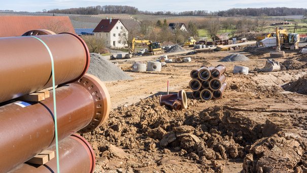 Im Vordergrund sieht man Wasserrohre, die in einem Neubaugebiet verlegt werden, im Hintergrund ein älterer Wohnhaus