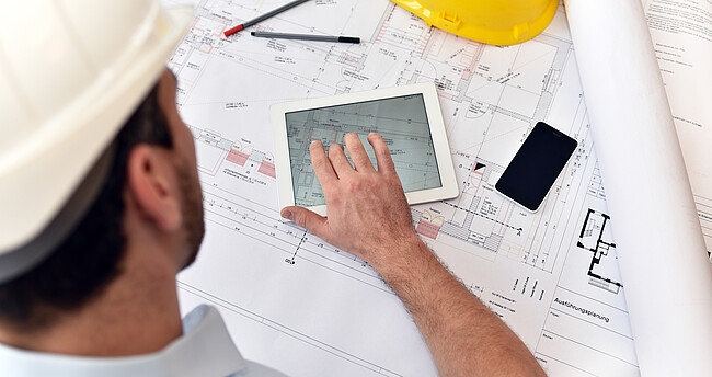 Ein Bauzeichner sitzt mit Tablet, handy und Zeichenutensilien an einem Plan