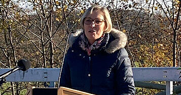 Eine Frau mit Brille steht auf einer Brücke hinter einem Rednerpult mit Mikrofon. 