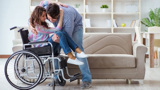 Bild zeigt junge Frau, die von einem Mann aus dem Rollstuhl gehoben wird