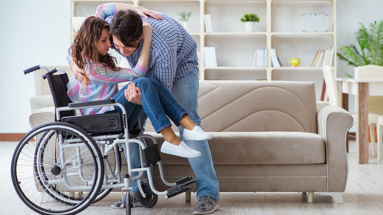 Bild zeigt junge Frau, die von einem Mann aus dem Rollstuhl gehoben wird