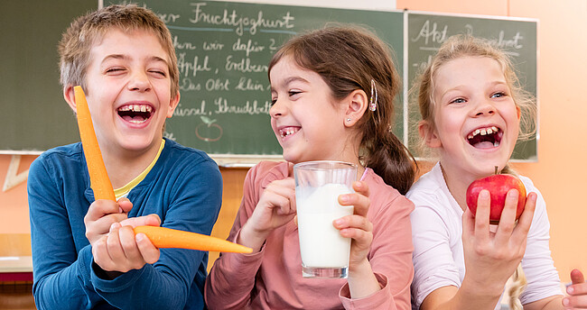 Eine Junge und zwei Mädchen sitzen nebeneinander und lachen in die Kamera, der junge spielt mit zwei Karotten, das Mädchen in der Mitte hält einen Apfel und das Mädchen daneben ein Glas Milch in der Hand