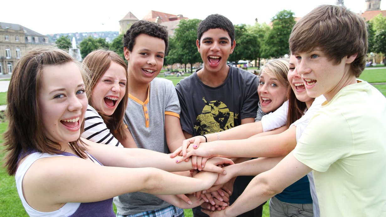 Jugendliche Mädchen und Jungs fassen sich an den Händen und lachen in die Kamera