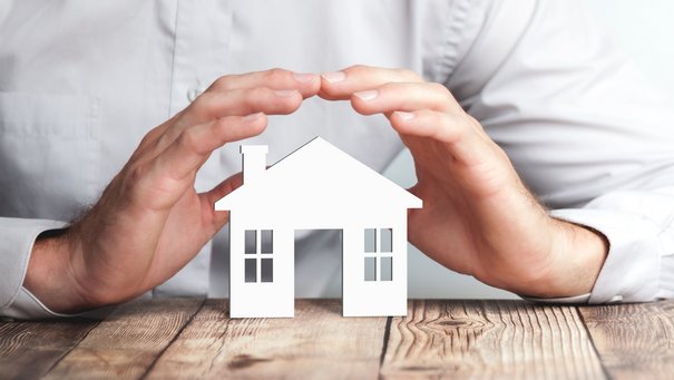 Eine männliche Hand hält seine Hände schützend über ein Miniaturhaus