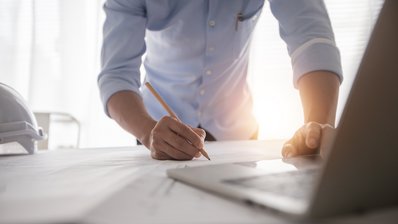 Ingenieur beugt sich über einen Plan und hat einen Stift in der Hand, im Vordergurnd sieht man einen Laptop