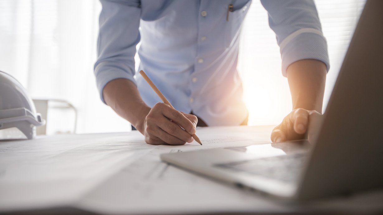 Ingenieur beugt sich über einen Plan und hat einen Stift in der Hand, im Vordergurnd sieht man einen Laptop