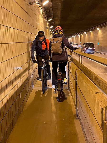 Zwei Fahrradfahrer begegnen sich auf einem sehr schmalen Fahrradweg im Flughafentunnel.